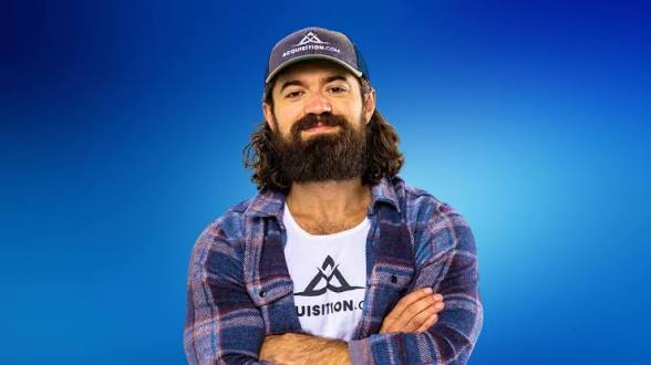 Portrait of Alex Hormozi in a plaid shirt and Acquisition.com cap against a blue backdrop.