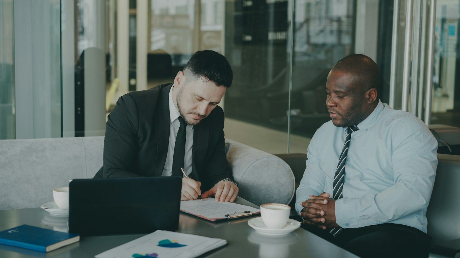 Three people sit at a bright meeting table discussing paperwork in natural daylight.